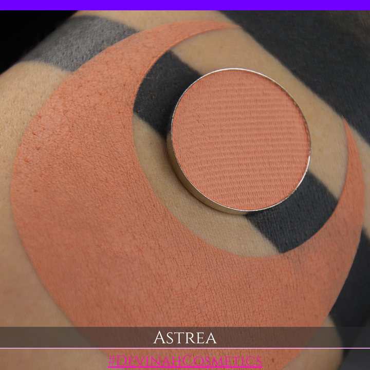 A close-up of a person's arm with a large peach-toned makeup swatch and a matching round eyeshadow makeup pan labeled ASTREA, with the text #DEVINAHCOSMETICS at the bottom.