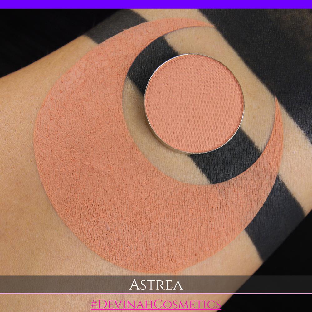 A close-up of a person's arm with a large peach-toned makeup swatch and a matching round eyeshadow makeup pan labeled ASTREA, with the text #DEVINAHCOSMETICS at the bottom.