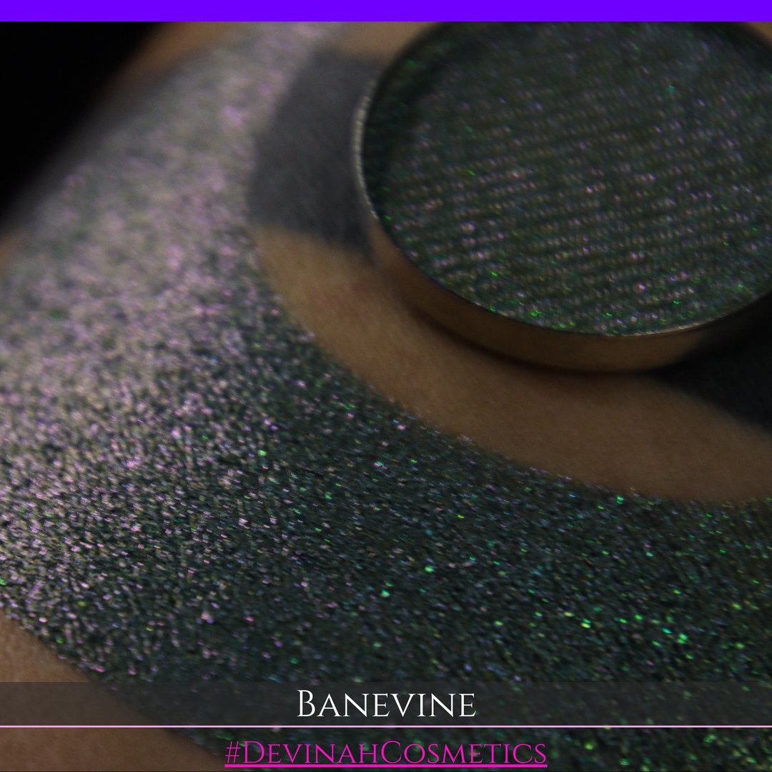 A close-up of a person's arm with a large crescent-shaped swatch of shimmering green eyeshadow, featuring a small round pan of the same green makeup placed on top. The text 'BANEVINE' and '#DEVINAHCOSMETICS' appear at the bottom.