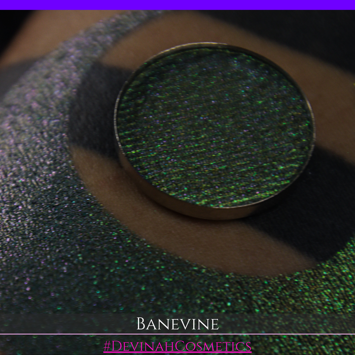 A close-up of a person's arm with a large crescent-shaped swatch of shimmering green eyeshadow, featuring a small round pan of the same green makeup placed on top. The text 'BANEVINE' and '#DEVINAHCOSMETICS' appear at the bottom.