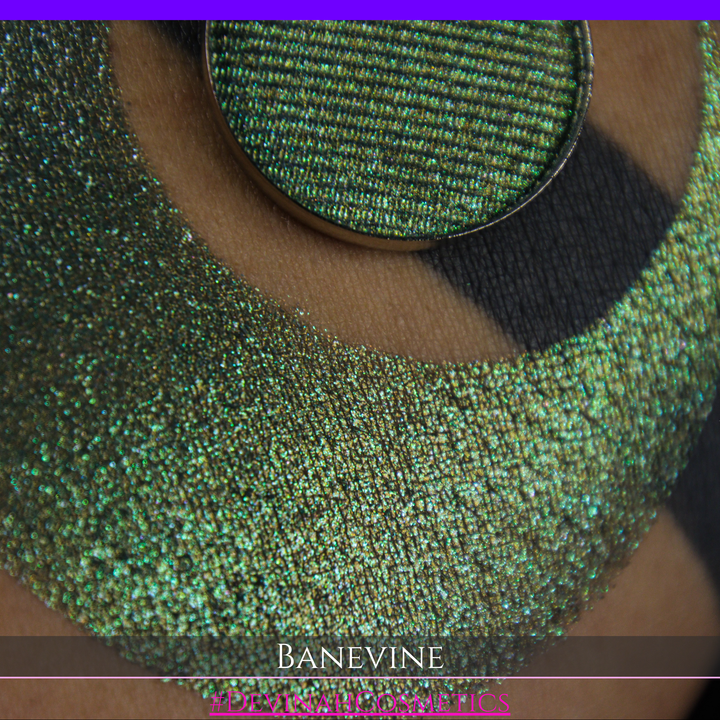 A close-up of a person's arm with a large crescent-shaped swatch of shimmering green eyeshadow, featuring a small round pan of the same green makeup placed on top. The text 'BANEVINE' and '#DEVINAHCOSMETICS' appear at the bottom.