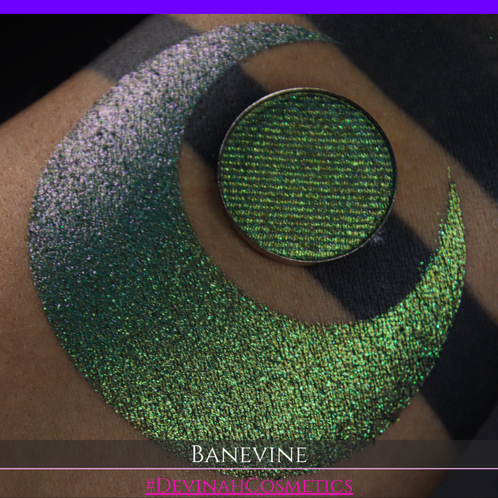 A close-up of a person's arm with a large crescent-shaped swatch of shimmering green eyeshadow, featuring a small round pan of the same green makeup placed on top. The text 'BANEVINE' and '#DEVINAHCOSMETICS' appear at the bottom.