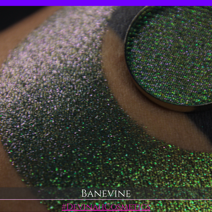 A close-up of a person's arm with a large crescent-shaped swatch of shimmering green eyeshadow, featuring a small round pan of the same green makeup placed on top. The text 'BANEVINE' and '#DEVINAHCOSMETICS' appear at the bottom.