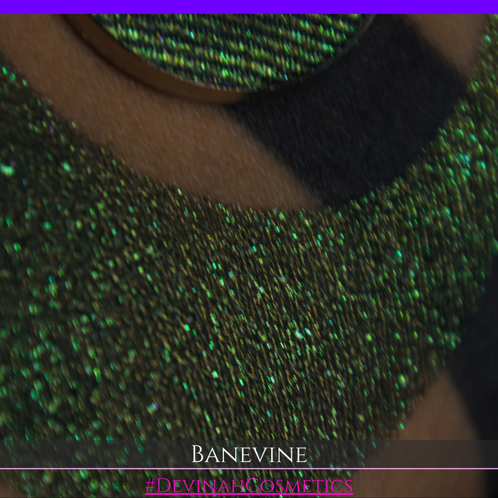A close-up of a person's arm with a large crescent-shaped swatch of shimmering green eyeshadow, featuring a small round pan of the same green makeup placed on top. The text 'BANEVINE' and '#DEVINAHCOSMETICS' appear at the bottom.