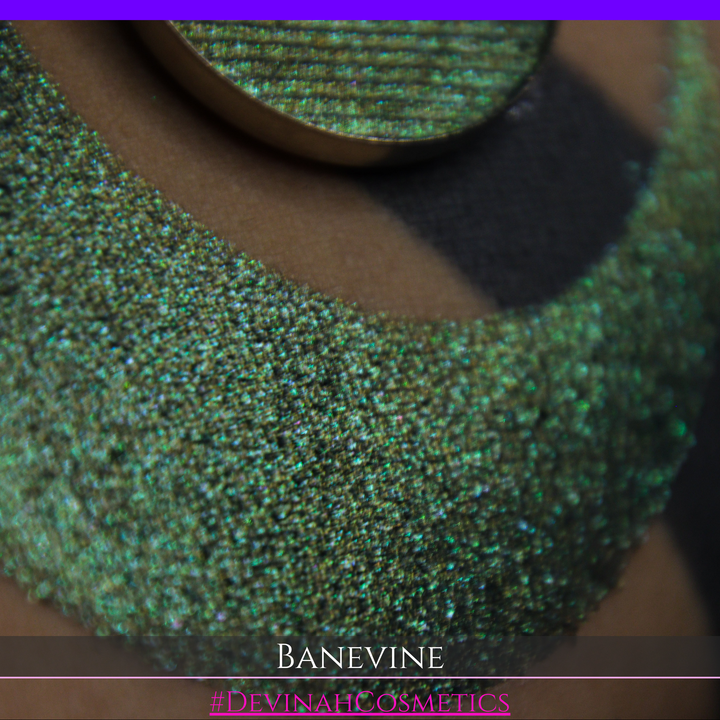 A close-up of a person's arm with a large crescent-shaped swatch of shimmering green eyeshadow, featuring a small round pan of the same green makeup placed on top. The text 'BANEVINE' and '#DEVINAHCOSMETICS' appear at the bottom.