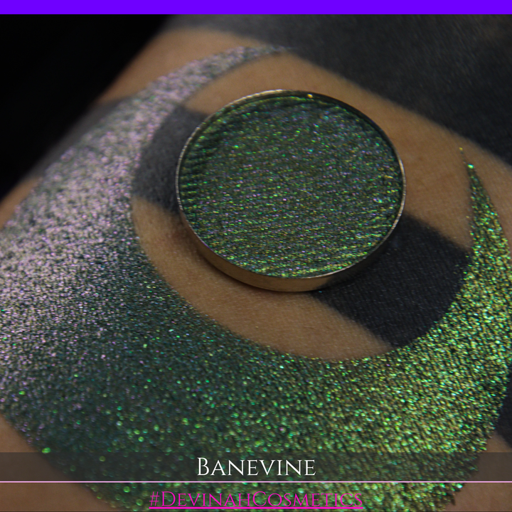 A close-up of a person's arm with a large crescent-shaped swatch of shimmering green eyeshadow, featuring a small round pan of the same green makeup placed on top. The text 'BANEVINE' and '#DEVINAHCOSMETICS' appear at the bottom.