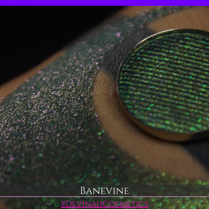A close-up of a person's arm with a large crescent-shaped swatch of shimmering green eyeshadow, featuring a small round pan of the same green makeup placed on top. The text 'BANEVINE' and '#DEVINAHCOSMETICS' appear at the bottom.