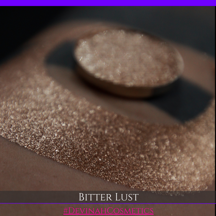 A round eyeshadow pan labeled 'BITTER LUST' made by Devinah Cosmetics, placed over a large crescent moon swatch of the same shimmering shifty eyeshadow on skin. The eyeshadow has shimmer sparkle colors brown, tan, beige, neutral.