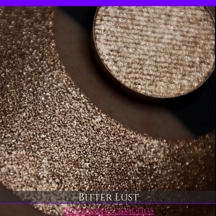 A round eyeshadow pan labeled 'BITTER LUST' made by Devinah Cosmetics, placed over a large crescent moon swatch of the same shimmering shifty eyeshadow on skin. The eyeshadow has shimmer sparkle colors brown, tan, beige, neutral.