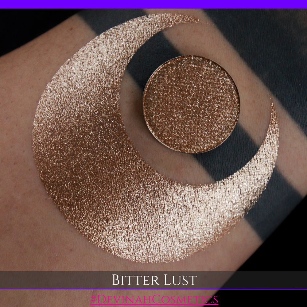 A round eyeshadow pan labeled 'BITTER LUST' made by Devinah Cosmetics, placed over a large crescent moon swatch of the same shimmering shifty eyeshadow on skin. The eyeshadow has shimmer sparkle colors brown, tan, beige, neutral.