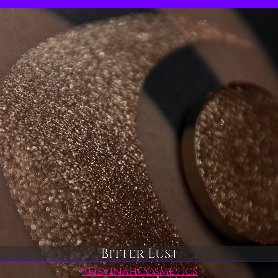 A round eyeshadow pan labeled 'BITTER LUST' made by Devinah Cosmetics, placed over a large crescent moon swatch of the same shimmering shifty eyeshadow on skin. The eyeshadow has shimmer sparkle colors brown, tan, beige, neutral.