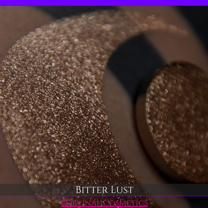 A round eyeshadow pan labeled 'BITTER LUST' made by Devinah Cosmetics, placed over a large crescent moon swatch of the same shimmering shifty eyeshadow on skin. The eyeshadow has shimmer sparkle colors brown, tan, beige, neutral.