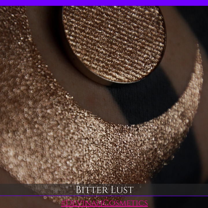 A round eyeshadow pan labeled 'BITTER LUST' made by Devinah Cosmetics, placed over a large crescent moon swatch of the same shimmering shifty eyeshadow on skin. The eyeshadow has shimmer sparkle colors brown, tan, beige, neutral.