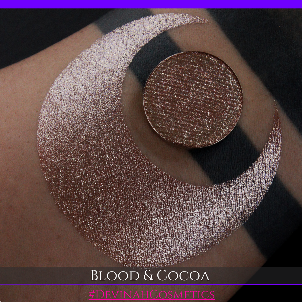 A round eyeshadow pan labeled 'BLOOD AND COCOA' made by Devinah Cosmetics, placed over a large crescent moon swatch of the same shimmering shifty eyeshadow on skin. The eyeshadow has shimmery sparkle of red infused chocolate shimmer.