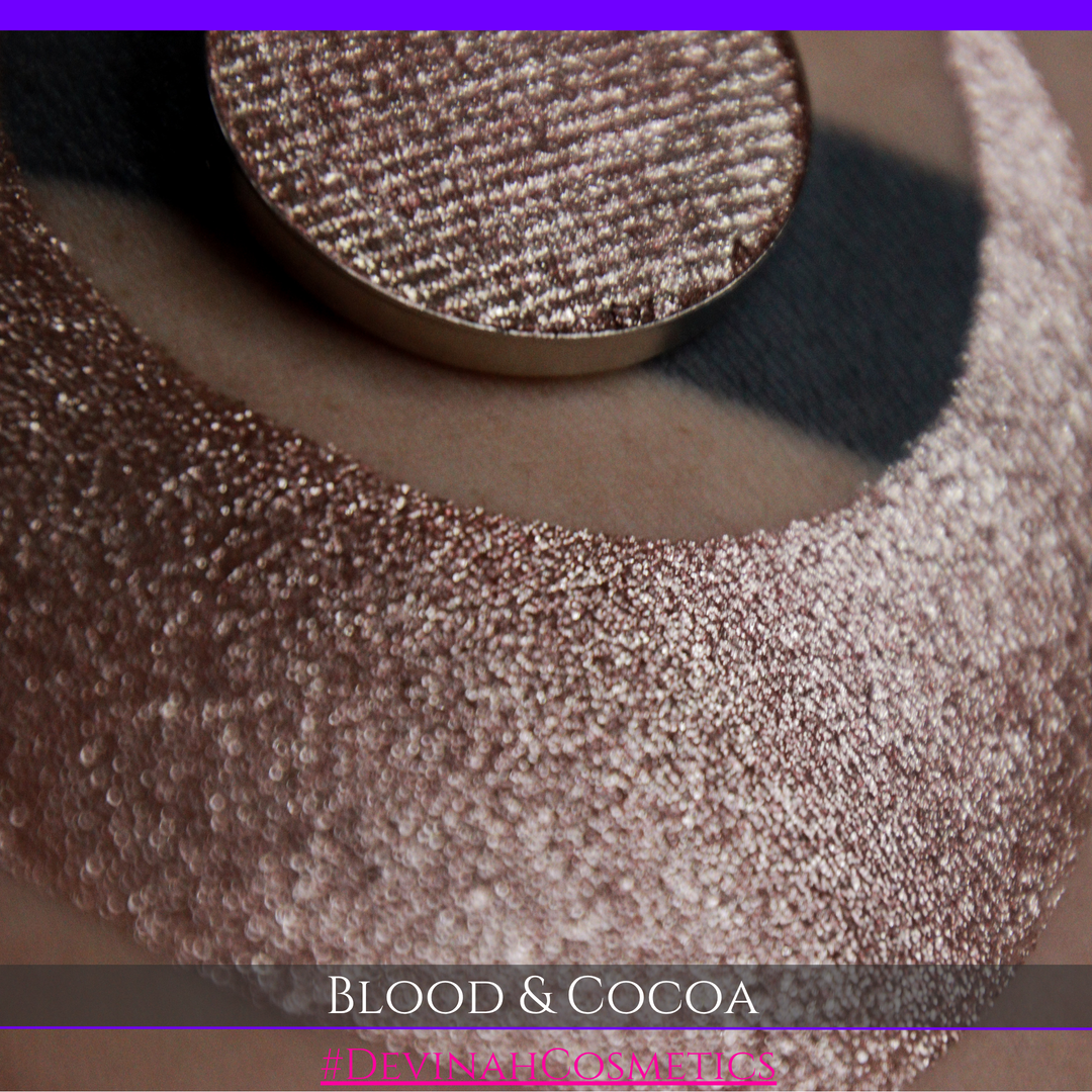 A round eyeshadow pan labeled 'BLOOD AND COCOA' made by Devinah Cosmetics, placed over a large crescent moon swatch of the same shimmering shifty eyeshadow on skin. The eyeshadow has shimmery sparkle of red infused chocolate shimmer.