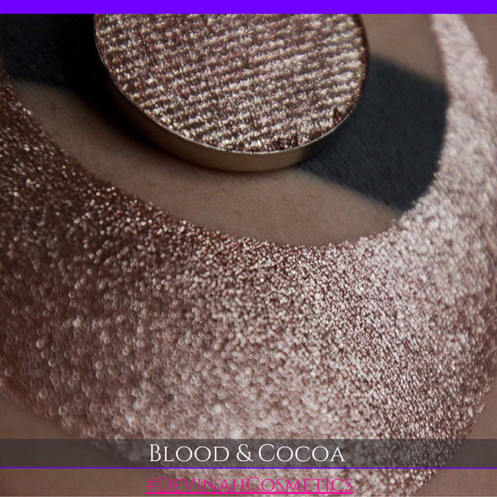A round eyeshadow pan labeled 'BLOOD AND COCOA' made by Devinah Cosmetics, placed over a large crescent moon swatch of the same shimmering shifty eyeshadow on skin. The eyeshadow has shimmery sparkle of red infused chocolate shimmer.