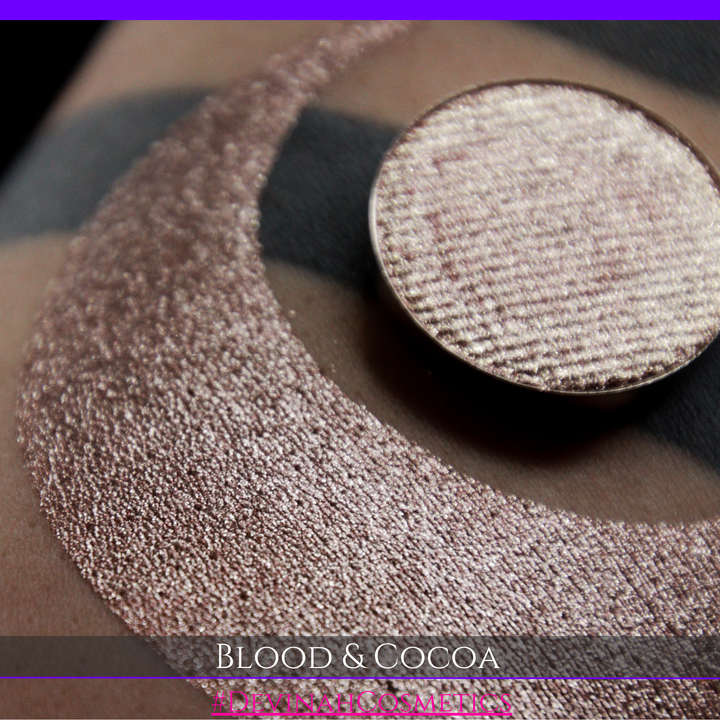 A round eyeshadow pan labeled 'BLOOD AND COCOA' made by Devinah Cosmetics, placed over a large crescent moon swatch of the same shimmering shifty eyeshadow on skin. The eyeshadow has shimmery sparkle of red infused chocolate shimmer.