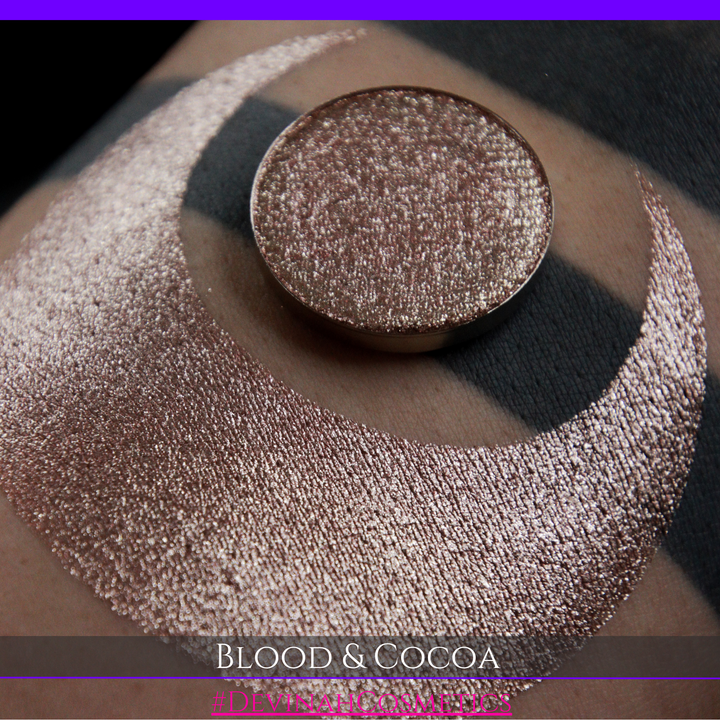 A round eyeshadow pan labeled 'BLOOD AND COCOA' made by Devinah Cosmetics, placed over a large crescent moon swatch of the same shimmering shifty eyeshadow on skin. The eyeshadow has shimmery sparkle of red infused chocolate shimmer.