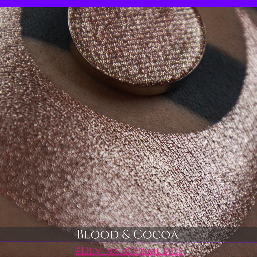 A round eyeshadow pan labeled 'BLOOD AND COCOA' made by Devinah Cosmetics, placed over a large crescent moon swatch of the same shimmering shifty eyeshadow on skin. The eyeshadow has shimmery sparkle of red infused chocolate shimmer.