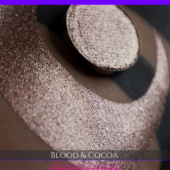A round eyeshadow pan labeled 'BLOOD AND COCOA' made by Devinah Cosmetics, placed over a large crescent moon swatch of the same shimmering shifty eyeshadow on skin. The eyeshadow has shimmery sparkle of red infused chocolate shimmer.