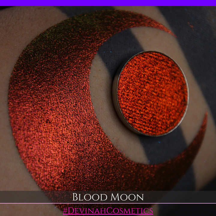 A close-up of a makeup swatch or eyeshadow pan labeled 'Blood Moon' showing a metallic, iridescent gradient from gold to orange to deep red, applied on skin with a matching circular eyeshadow pan above it. The text '#DevinahCosmetics' is visible at the bottom.