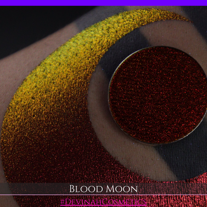 A close-up of a makeup swatch or eyeshadow pan labeled 'Blood Moon' showing a metallic, iridescent gradient from gold to orange to deep red, applied on skin with a matching circular eyeshadow pan above it. The text '#DevinahCosmetics' is visible at the bottom.