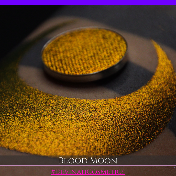 A close-up of a makeup swatch or eyeshadow pan labeled 'Blood Moon' showing a metallic, iridescent gradient from gold to orange to deep red, applied on skin with a matching circular eyeshadow pan above it. The text '#DevinahCosmetics' is visible at the bottom.