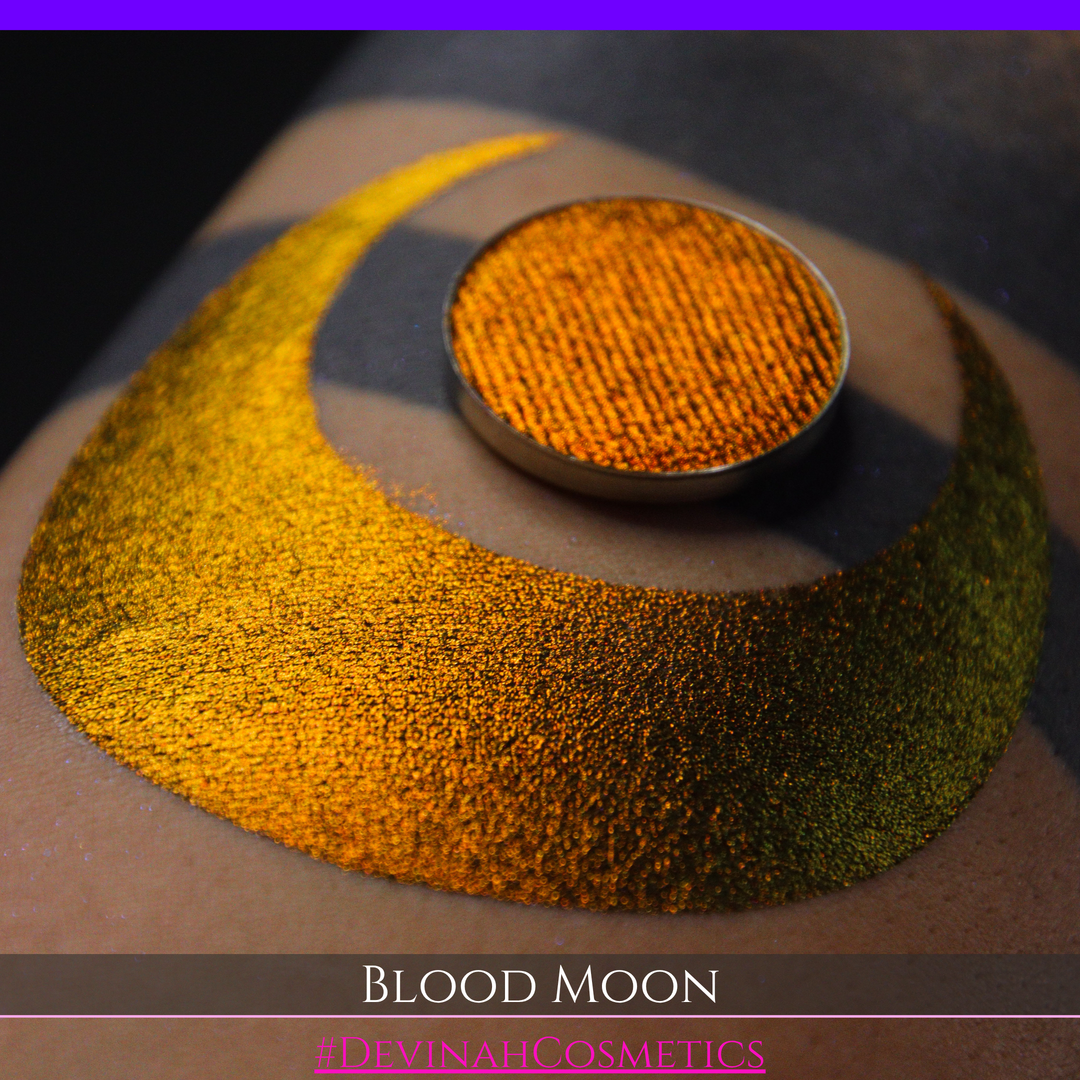 A close-up of a makeup swatch or eyeshadow pan labeled 'Blood Moon' showing a metallic, iridescent gradient from gold to orange to deep red, applied on skin with a matching circular eyeshadow pan above it. The text '#DevinahCosmetics' is visible at the bottom.