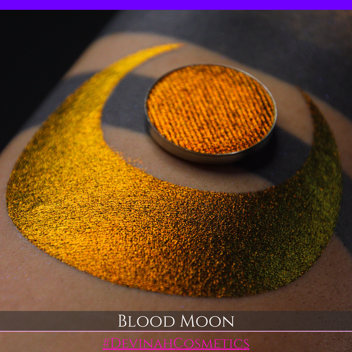 A close-up of a makeup swatch or eyeshadow pan labeled 'Blood Moon' showing a metallic, iridescent gradient from gold to orange to deep red, applied on skin with a matching circular eyeshadow pan above it. The text '#DevinahCosmetics' is visible at the bottom.