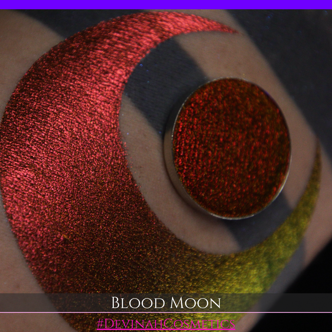 A close-up of a makeup swatch or eyeshadow pan labeled 'Blood Moon' showing a metallic, iridescent gradient from gold to orange to deep red, applied on skin with a matching circular eyeshadow pan above it. The text '#DevinahCosmetics' is visible at the bottom.