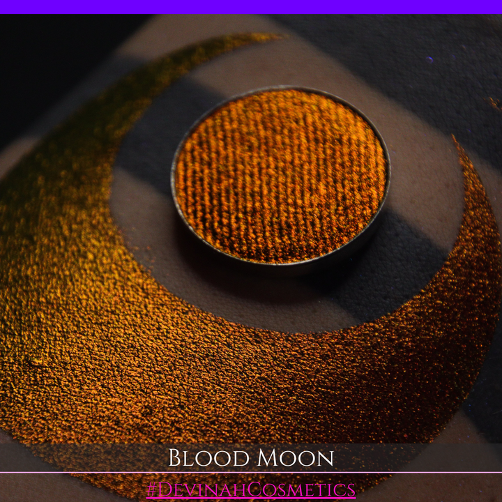 A close-up of a makeup swatch or eyeshadow pan labeled 'Blood Moon' showing a metallic, iridescent gradient from gold to orange to deep red, applied on skin with a matching circular eyeshadow pan above it. The text '#DevinahCosmetics' is visible at the bottom.