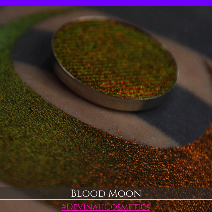 A close-up of a makeup swatch or eyeshadow pan labeled 'Blood Moon' showing a metallic, iridescent gradient from gold to orange to deep red, applied on skin with a matching circular eyeshadow pan above it. The text '#DevinahCosmetics' is visible at the bottom.