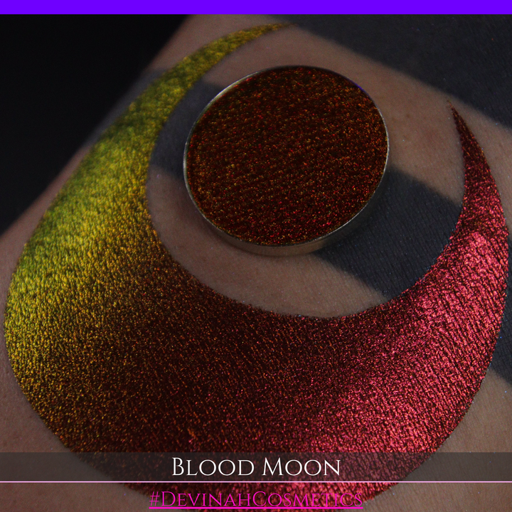 A close-up of a makeup swatch or eyeshadow pan labeled 'Blood Moon' showing a metallic, iridescent gradient from gold to orange to deep red, applied on skin with a matching circular eyeshadow pan above it. The text '#DevinahCosmetics' is visible at the bottom.