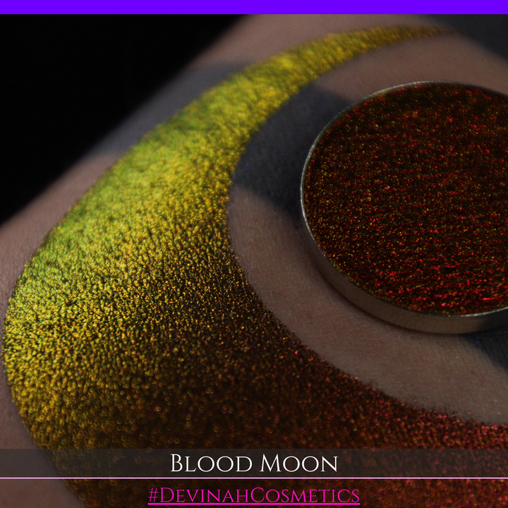 A close-up of a makeup swatch or eyeshadow pan labeled 'Blood Moon' showing a metallic, iridescent gradient from gold to orange to deep red, applied on skin with a matching circular eyeshadow pan above it. The text '#DevinahCosmetics' is visible at the bottom.