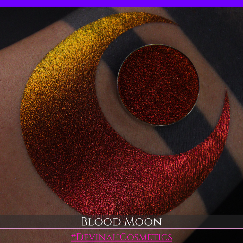 A close-up of a makeup swatch or eyeshadow pan labeled 'Blood Moon' showing a metallic, iridescent gradient from gold to orange to deep red, applied on skin with a matching circular eyeshadow pan above it. The text '#DevinahCosmetics' is visible at the bottom.