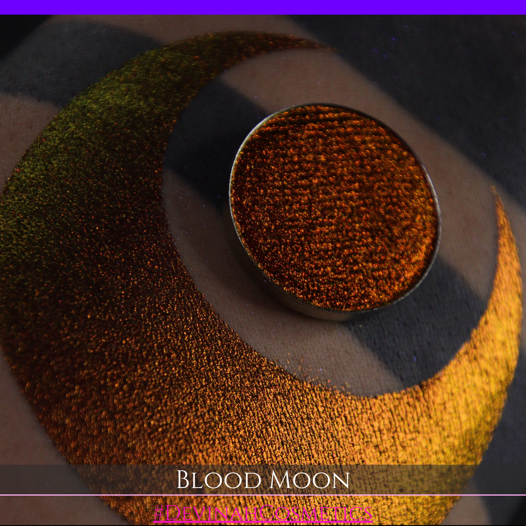 A close-up of a makeup swatch or eyeshadow pan labeled 'Blood Moon' showing a metallic, iridescent gradient from gold to orange to deep red, applied on skin with a matching circular eyeshadow pan above it. The text '#DevinahCosmetics' is visible at the bottom.