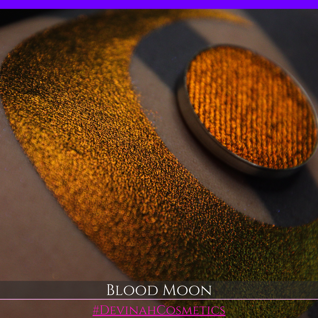 A close-up of a makeup swatch or eyeshadow pan labeled 'Blood Moon' showing a metallic, iridescent gradient from gold to orange to deep red, applied on skin with a matching circular eyeshadow pan above it. The text '#DevinahCosmetics' is visible at the bottom.