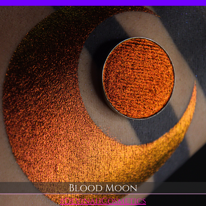 A close-up of a makeup swatch or eyeshadow pan labeled 'Blood Moon' showing a metallic, iridescent gradient from gold to orange to deep red, applied on skin with a matching circular eyeshadow pan above it. The text '#DevinahCosmetics' is visible at the bottom.