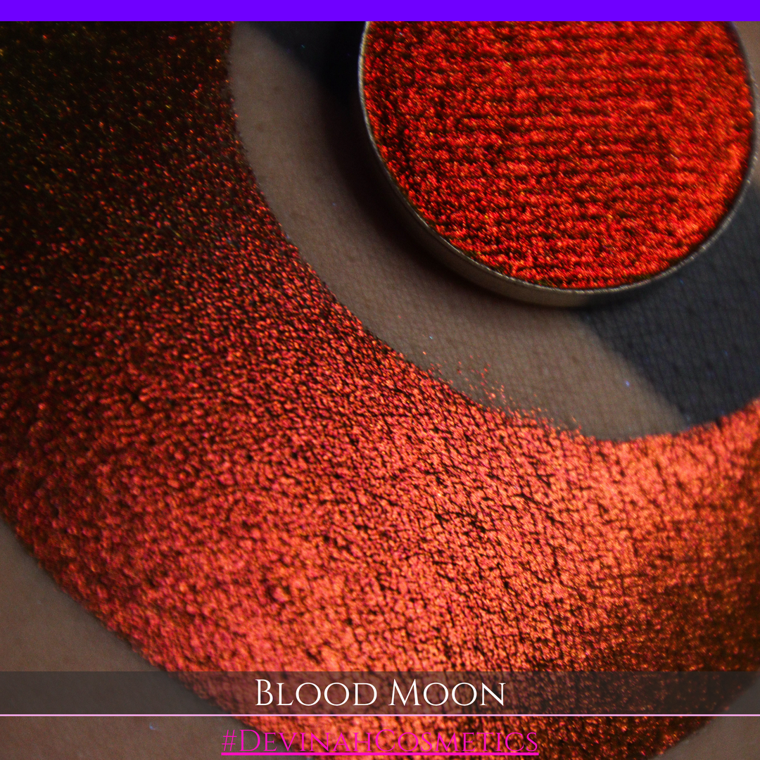 A close-up of a makeup swatch or eyeshadow pan labeled 'Blood Moon' showing a metallic, iridescent gradient from gold to orange to deep red, applied on skin with a matching circular eyeshadow pan above it. The text '#DevinahCosmetics' is visible at the bottom.