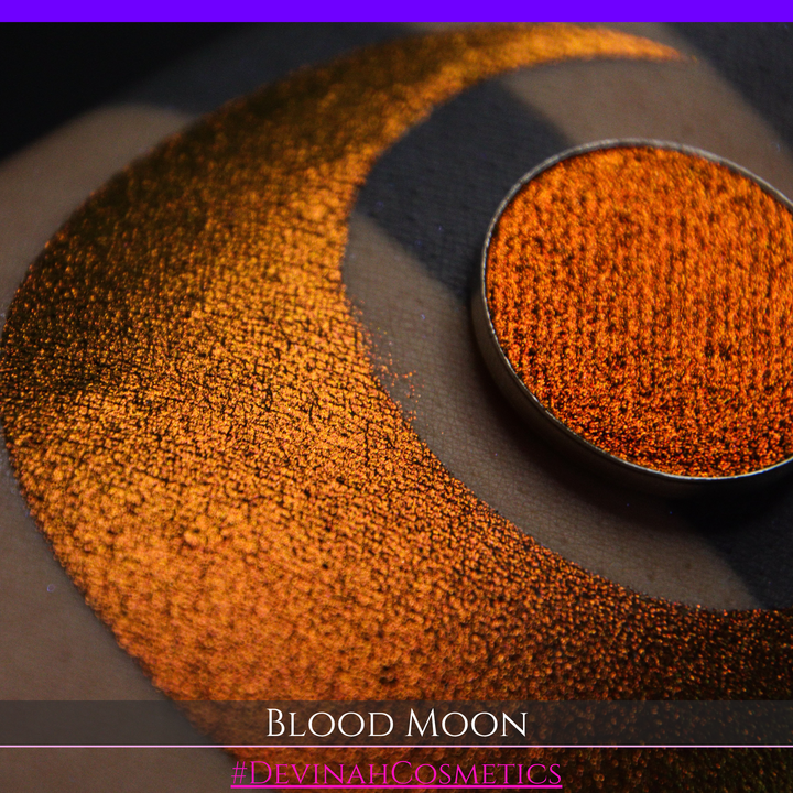 A close-up of a makeup swatch or eyeshadow pan labeled 'Blood Moon' showing a metallic, iridescent gradient from gold to orange to deep red, applied on skin with a matching circular eyeshadow pan above it. The text '#DevinahCosmetics' is visible at the bottom.