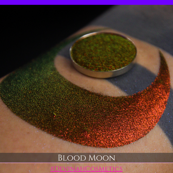 A close-up of a makeup swatch or eyeshadow pan labeled 'Blood Moon' showing a metallic, iridescent gradient from gold to orange to deep red, applied on skin with a matching circular eyeshadow pan above it. The text '#DevinahCosmetics' is visible at the bottom.