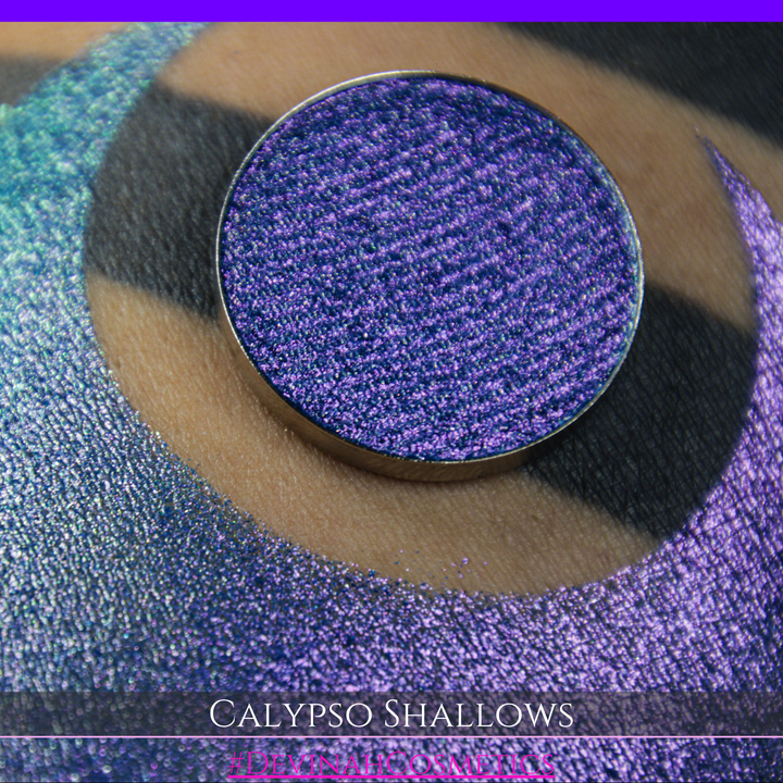 A round eyeshadow pan labeled 'CALYPSO SHALLOWS' made by Devinah Cosmetics, placed over a large crescent moon swatch of the same shimmering shifty eyeshadow on skin. The eyeshadow has a shift between colors gree, purple, blue.