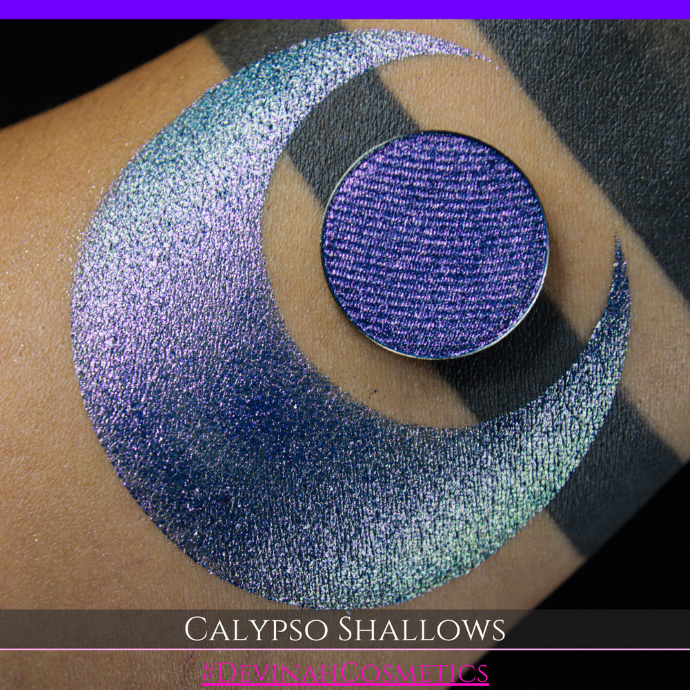 A round eyeshadow pan labeled 'CALYPSO SHALLOWS' made by Devinah Cosmetics, placed over a large crescent moon swatch of the same shimmering shifty eyeshadow on skin. The eyeshadow has a shift between colors gree, purple, blue.