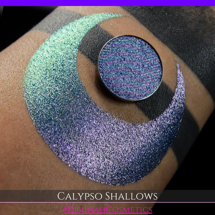 A round eyeshadow pan labeled 'CALYPSO SHALLOWS' made by Devinah Cosmetics, placed over a large crescent moon swatch of the same shimmering shifty eyeshadow on skin. The eyeshadow has a shift between colors gree, purple, blue.