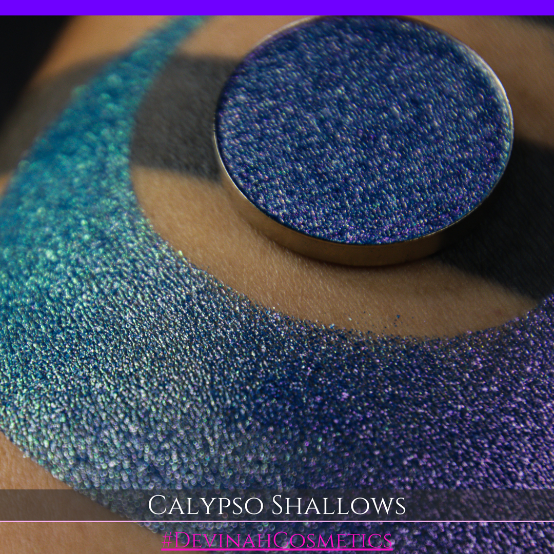 A round eyeshadow pan labeled 'CALYPSO SHALLOWS' made by Devinah Cosmetics, placed over a large crescent moon swatch of the same shimmering shifty eyeshadow on skin. The eyeshadow has a shift between colors gree, purple, blue.