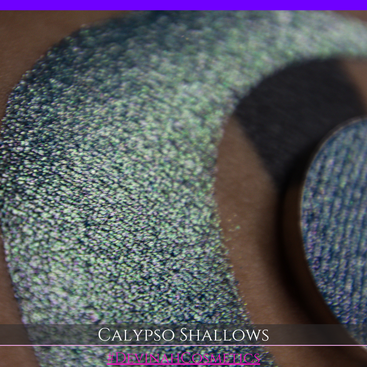 A round eyeshadow pan labeled 'CALYPSO SHALLOWS' made by Devinah Cosmetics, placed over a large crescent moon swatch of the same shimmering shifty eyeshadow on skin. The eyeshadow has a shift between colors gree, purple, blue.