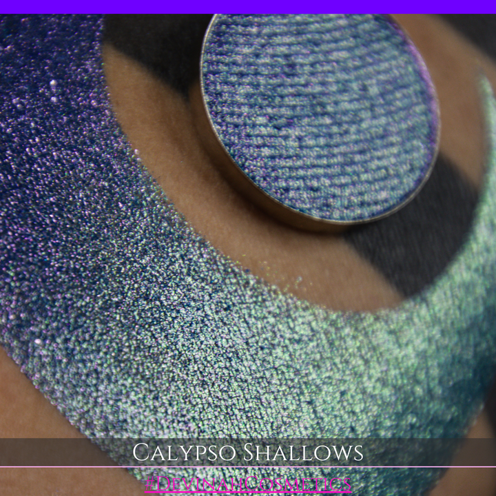 A round eyeshadow pan labeled 'CALYPSO SHALLOWS' made by Devinah Cosmetics, placed over a large crescent moon swatch of the same shimmering shifty eyeshadow on skin. The eyeshadow has a shift between colors gree, purple, blue.
