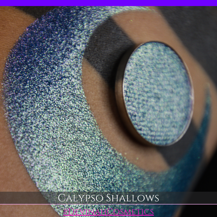 A round eyeshadow pan labeled 'CALYPSO SHALLOWS' made by Devinah Cosmetics, placed over a large crescent moon swatch of the same shimmering shifty eyeshadow on skin. The eyeshadow has a shift between colors gree, purple, blue.