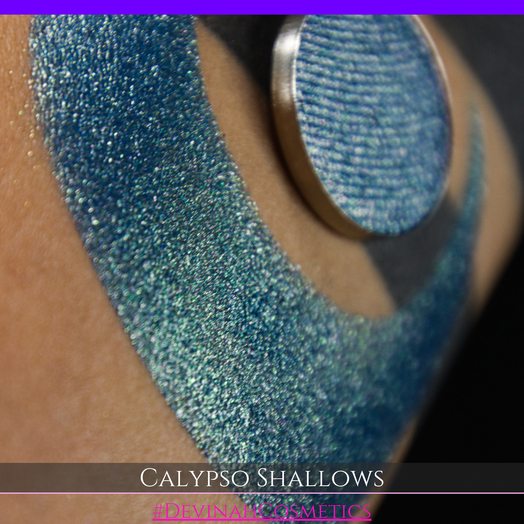 A round eyeshadow pan labeled 'CALYPSO SHALLOWS' made by Devinah Cosmetics, placed over a large crescent moon swatch of the same shimmering shifty eyeshadow on skin. The eyeshadow has a shift between colors gree, purple, blue.