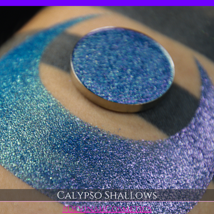 A round eyeshadow pan labeled 'CALYPSO SHALLOWS' made by Devinah Cosmetics, placed over a large crescent moon swatch of the same shimmering shifty eyeshadow on skin. The eyeshadow has a shift between colors gree, purple, blue.